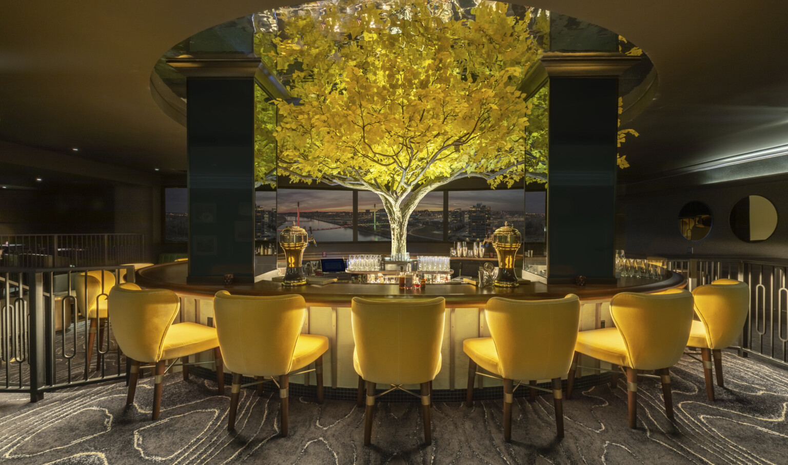 an illuminated yellow tree in the midst of in the round high top bar with yellow seats. Tree rises above round ceiling cutout