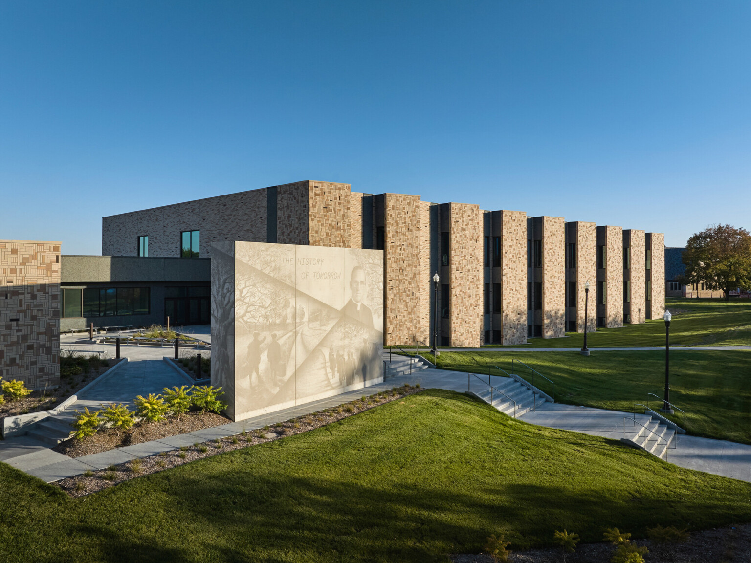 Boys Town Education Center exterior wall; red-tan stone with slated windows; shaded green space and walk paths in front of the wall; Stone structure with a mural light with phrase the history of tomorrow
