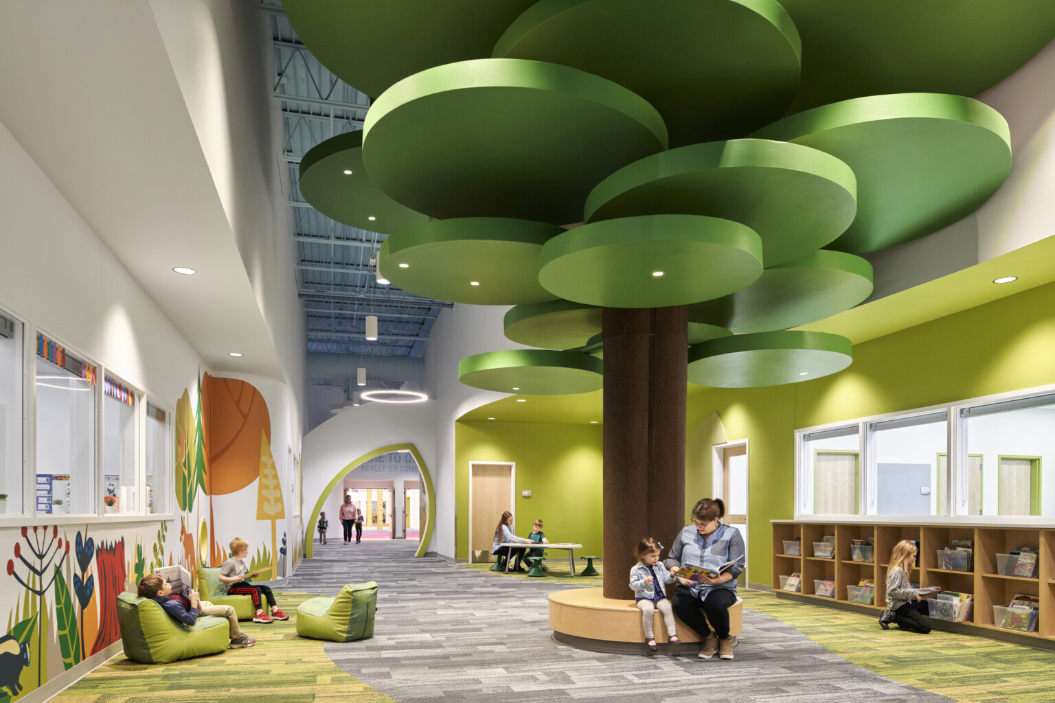 people reading on a brown circular bench with a brown column and green ceiling fixtures in hallway lined with forest graphics; dlr group design