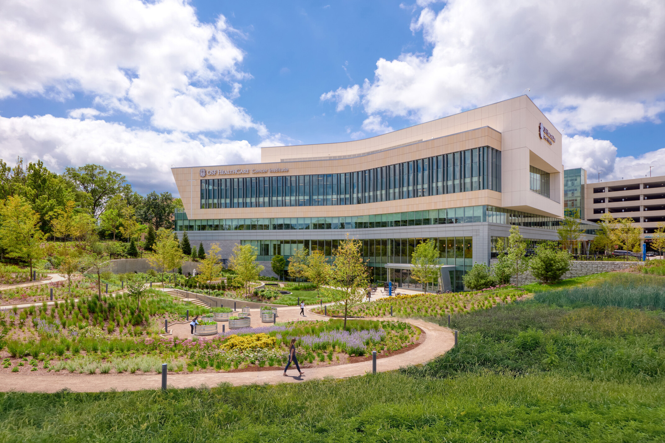 OSF Cancer Institute and Proton Therapy healthcare building; foreground greenery and walking space with the large modern building behind; featuring floor to ceiling windows facing out to the green space