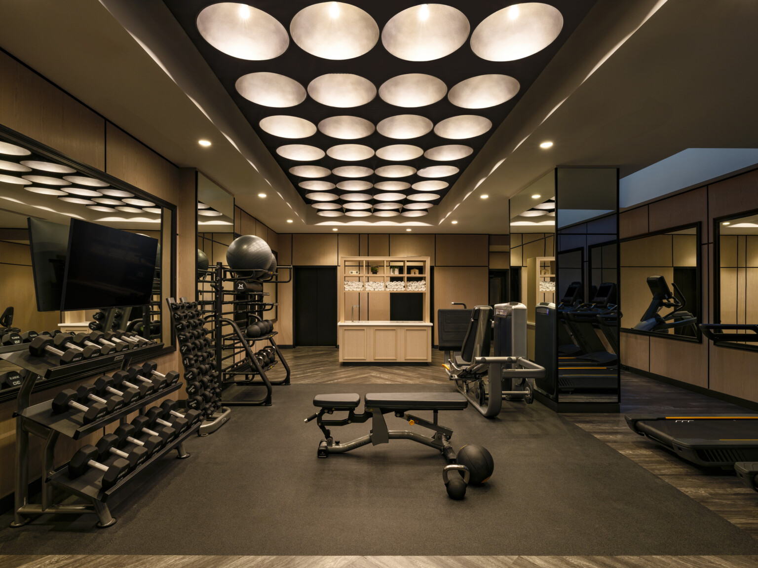 The Vanguard Hotel, Autograph Collection, gym space; circular light fixtures offer low light in the space; free weights and machines line the mirrored walls; the black machinery offsets the amber hue of the space