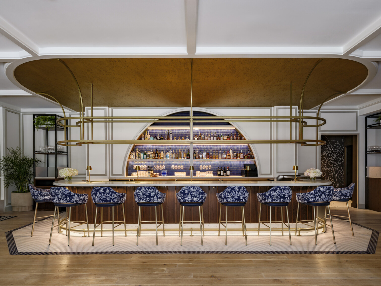 The Vanguard Hotel, Autograph Collection lobby bar area; medium wood and gold accented call and shelf features; blue high top chairs line the bar; an arched liquor wall in the wall behind the bar