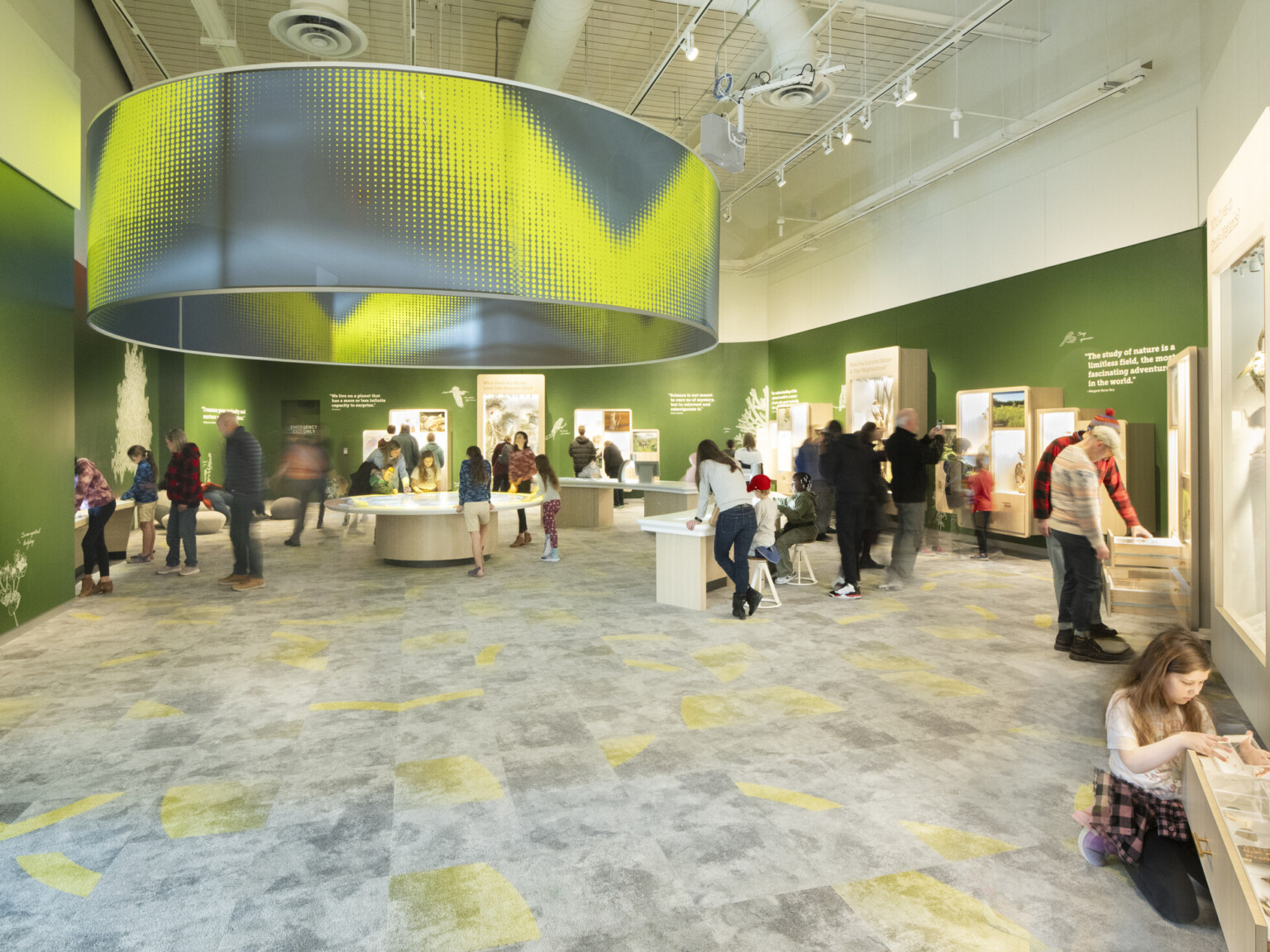 Visitors interacting with exhibits in a brightly lit indoor exhibition space with green walls and a large circular ceiling fixture