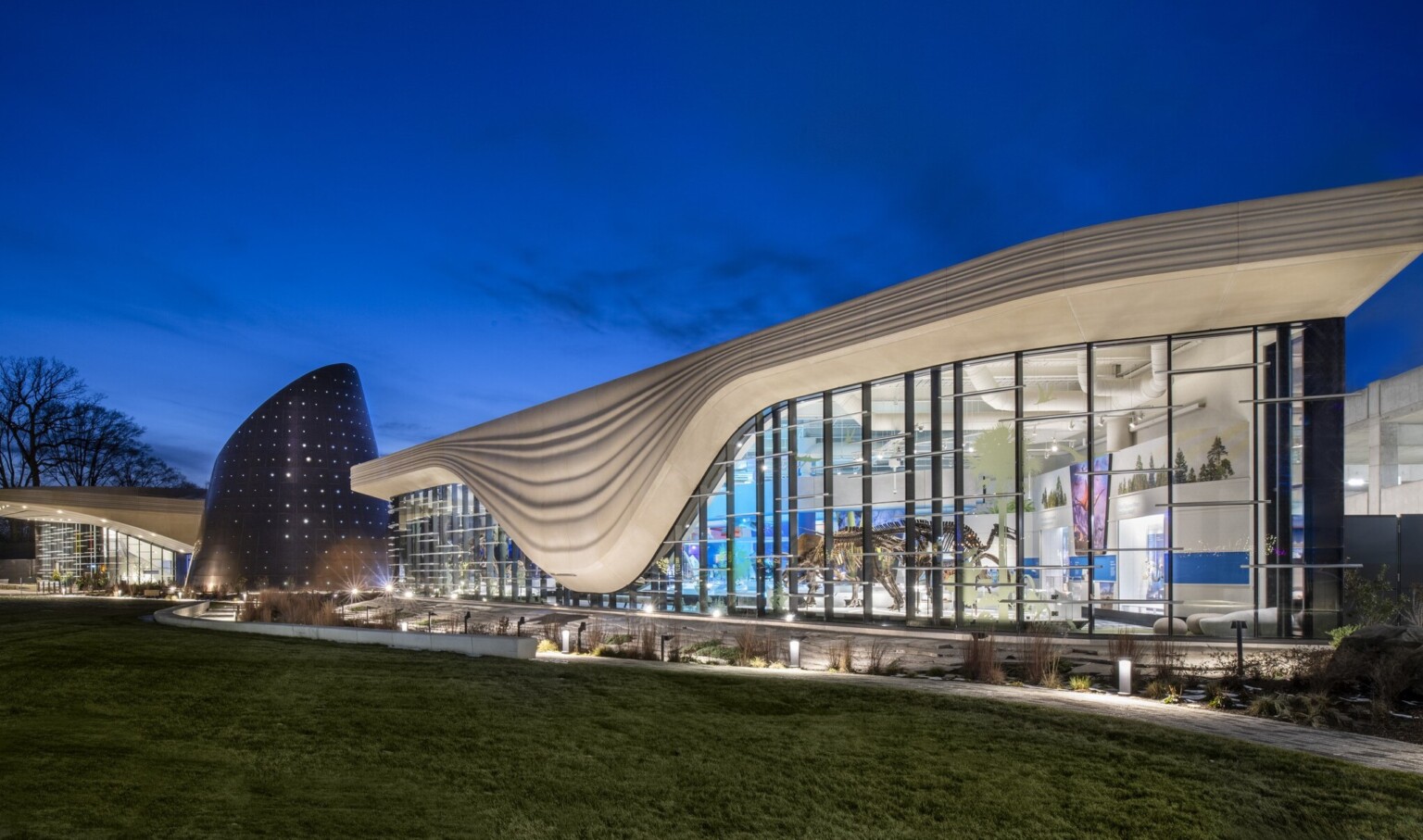 Nighttime image of a building with a white striped overhang that bows in center of right building section, an angled tube metal structure is at the building center
