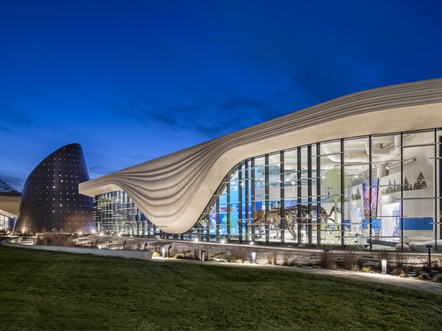 Nighttime image of a building with a white striped overhang that bows in center of right building section, an angled tube metal structure is at the building center