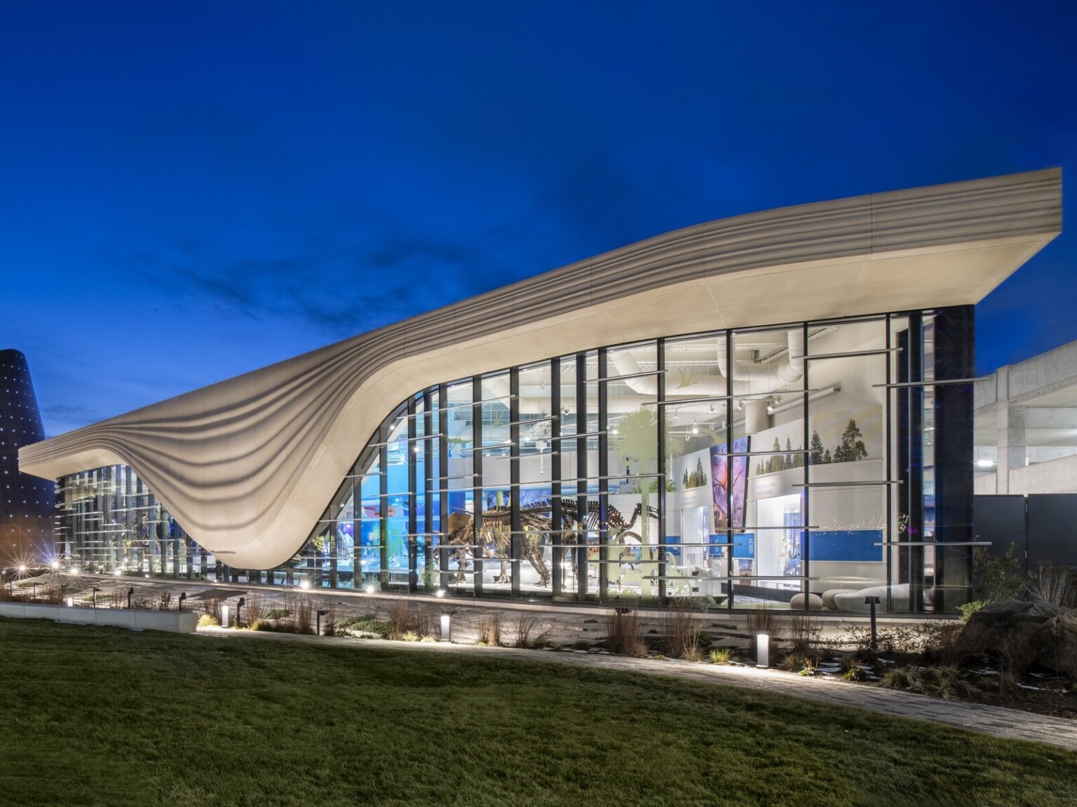 Nighttime image of a building with a white striped overhang that bows in center of right building section, an angled tube metal structure is at the building center