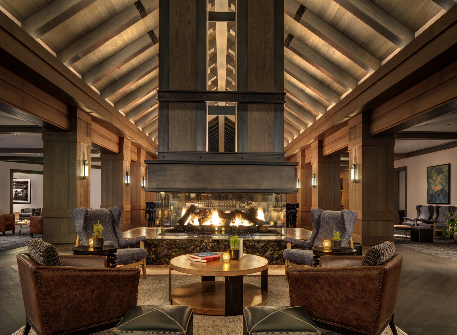 Cozy hotel lounge with a large double-sided fireplace, wooden ceiling beams and columns, and elegant seating arrangements throughout; dlr group design