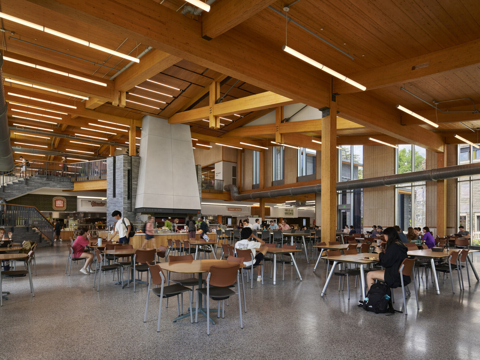 large open cafeteria with multiple storeys with seating for students; Mass timber ceiling makes for a warm and industrial space with exposed stone and venting