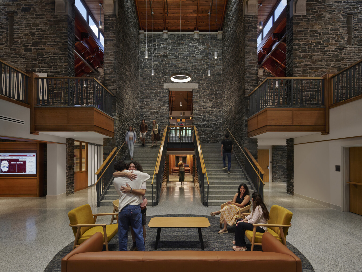 Broad staircase with wooden railings ascending to a second floor, flanked by stone walls, circular light fixture suspended from the ceiling. Seating area with yellow and brown chairs and a rectangular wooden coffee table on a speckled gray floor at the base of the stairs