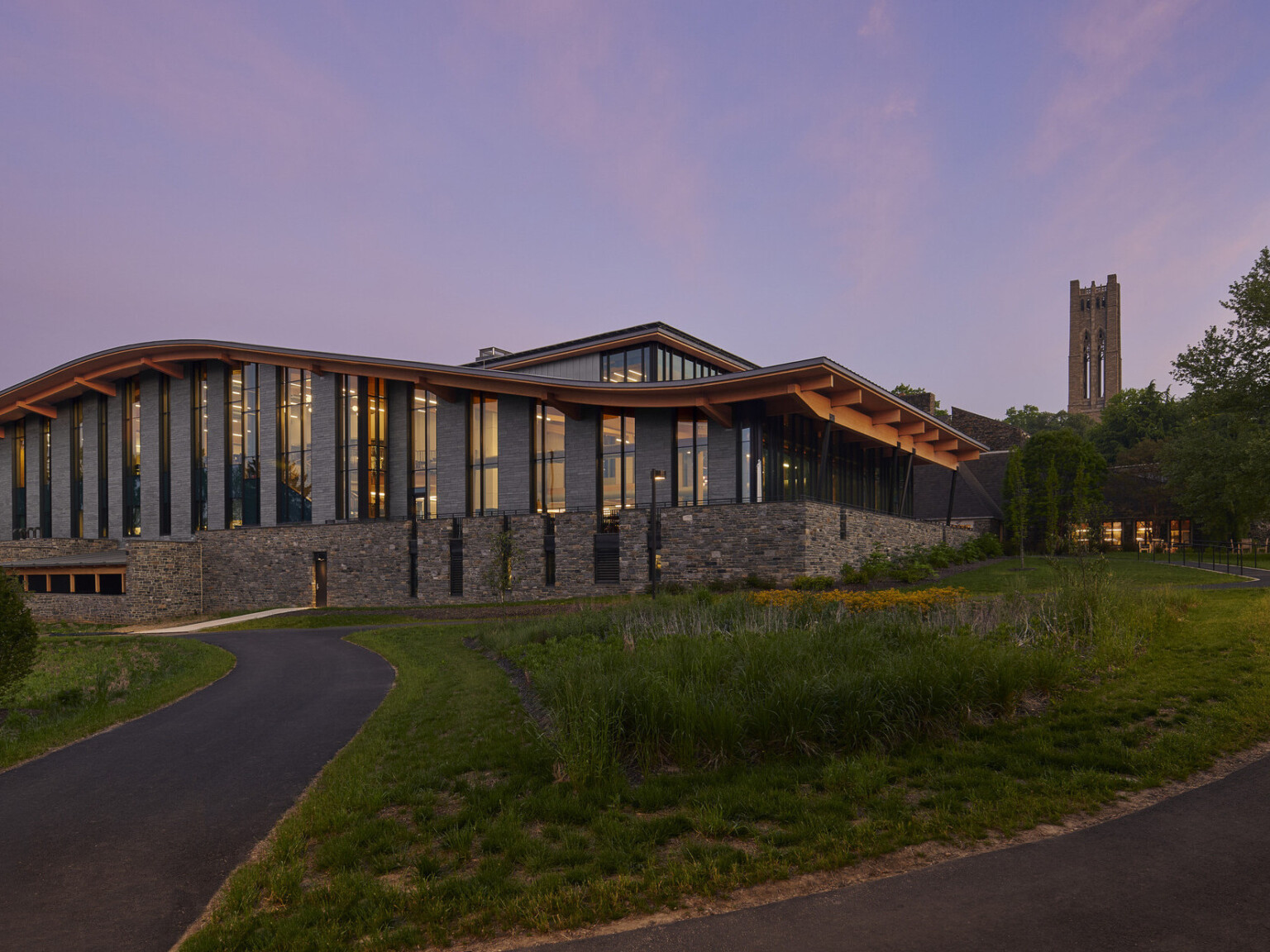 Stone and glass facade on university building with curving roof and overhang illuminated from within in the evening, mass timber architecture, what is mass timber construction