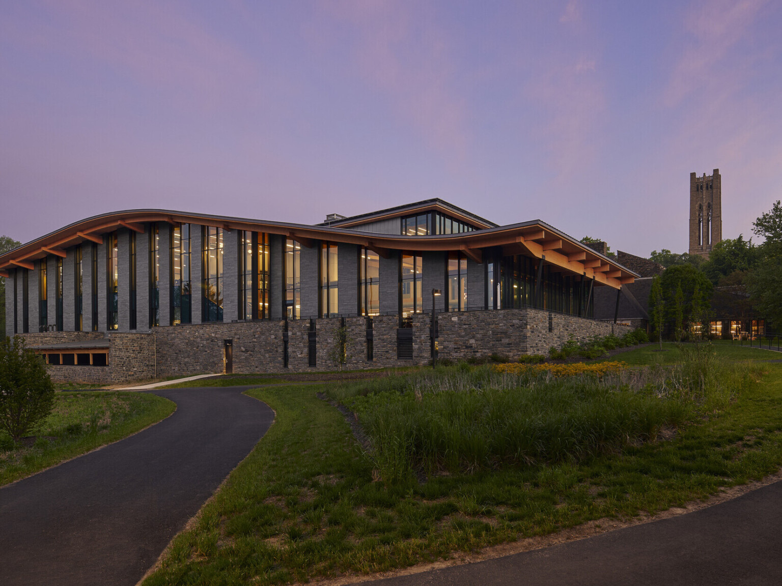 Stone and glass facade on university building with curving roof and overhang illuminated from within in the evening, mass timber architecture, what is mass timber construction