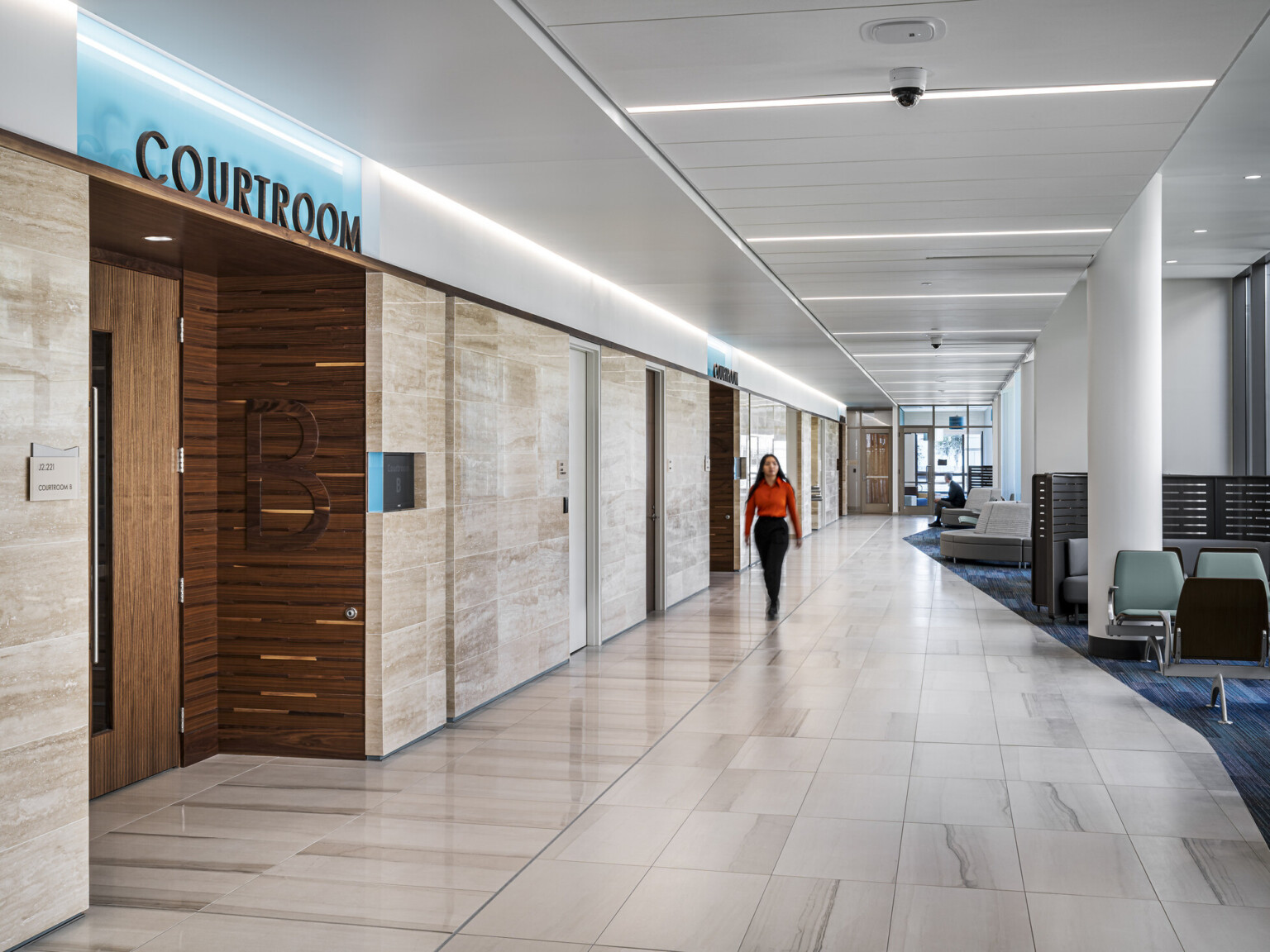 Woman dressed in a red long sleeve shirt walking down a hallway exterior to Courtroom B in the The Pinellas County Family Courts Annex; public seating and lounge space near windows.