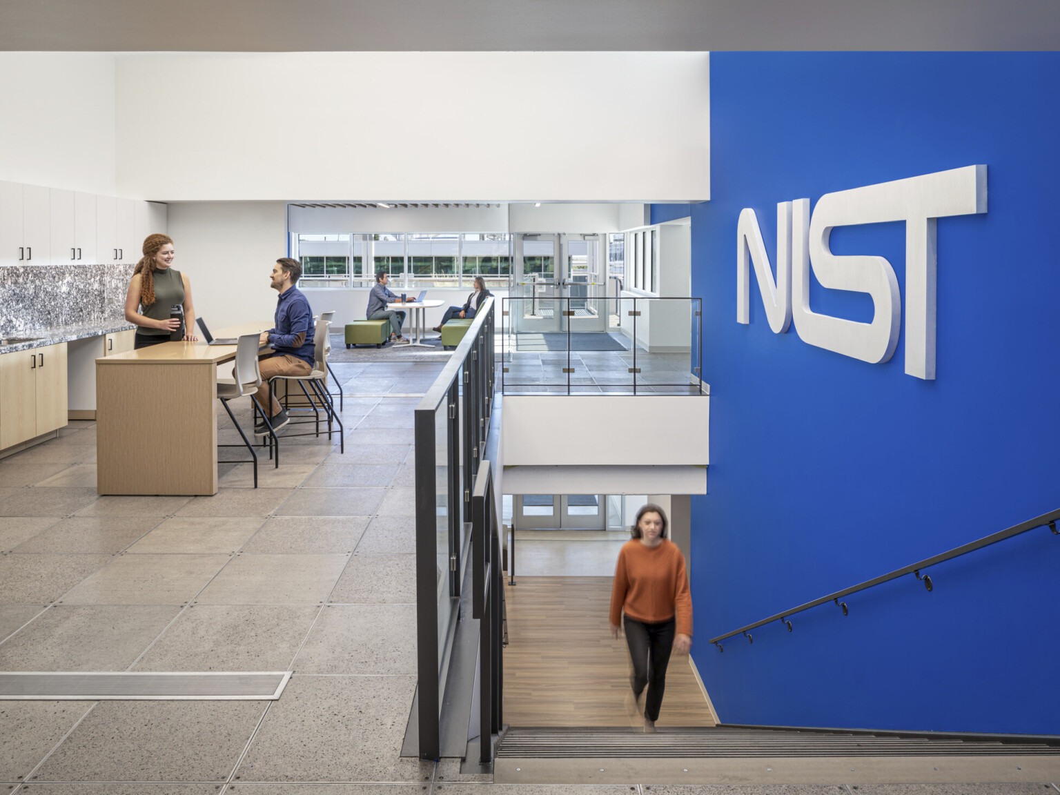 Woman in a orange shirt walking up a staircase with a blue wall to the right with letters NIST. Two couples at the top of the stairs working in collaborative areas; NIST Boulder