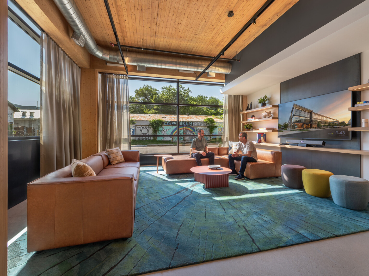Modern seating area filled with brown leather couches, rug in shades of blue, ottomans, walls with shelves and TV and a colorful mural outside the windows