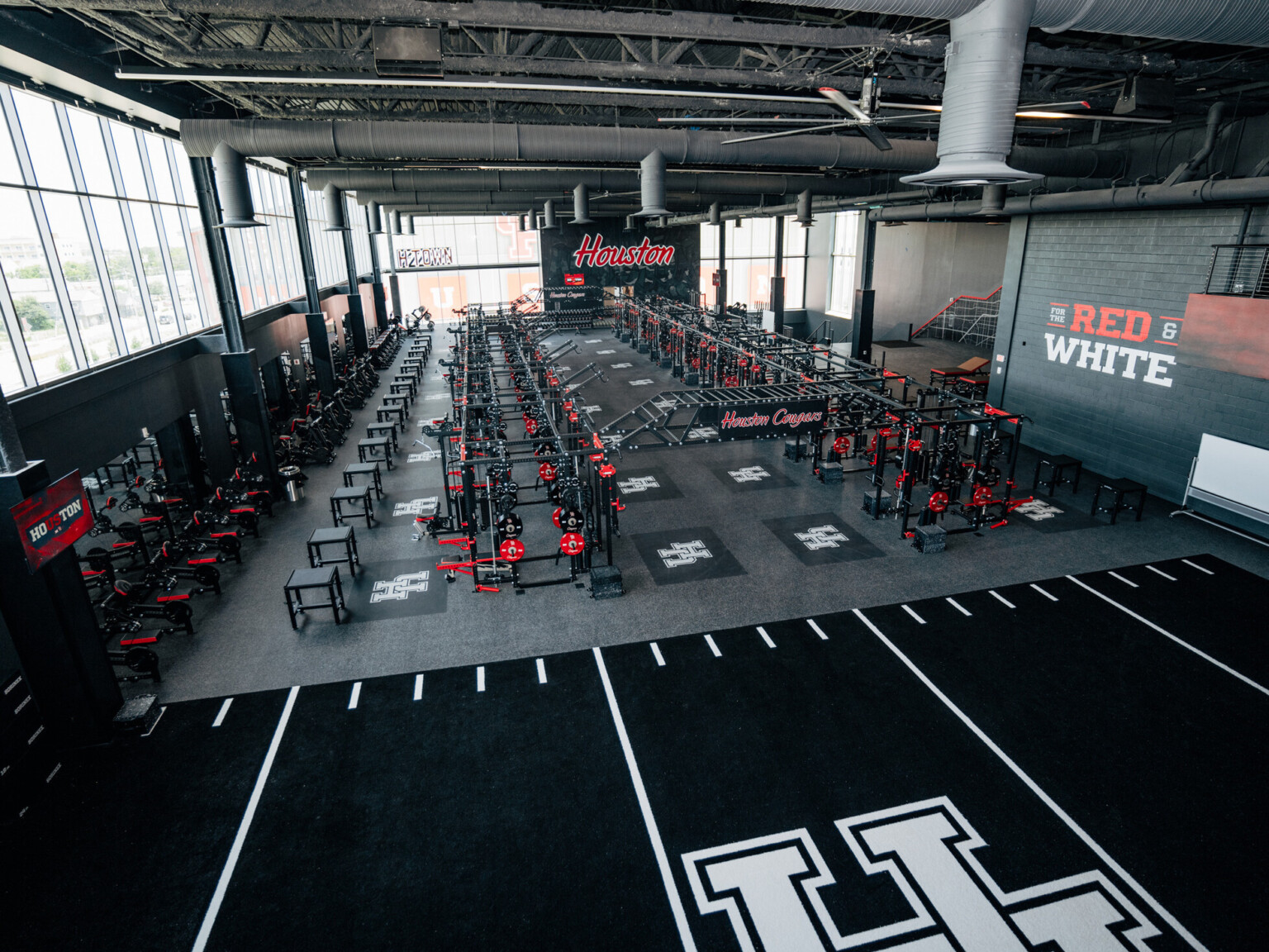 University of Houston football operations center weightroom with large UH logos on a indoor field space in front of 2 rows of weight lifting machinery