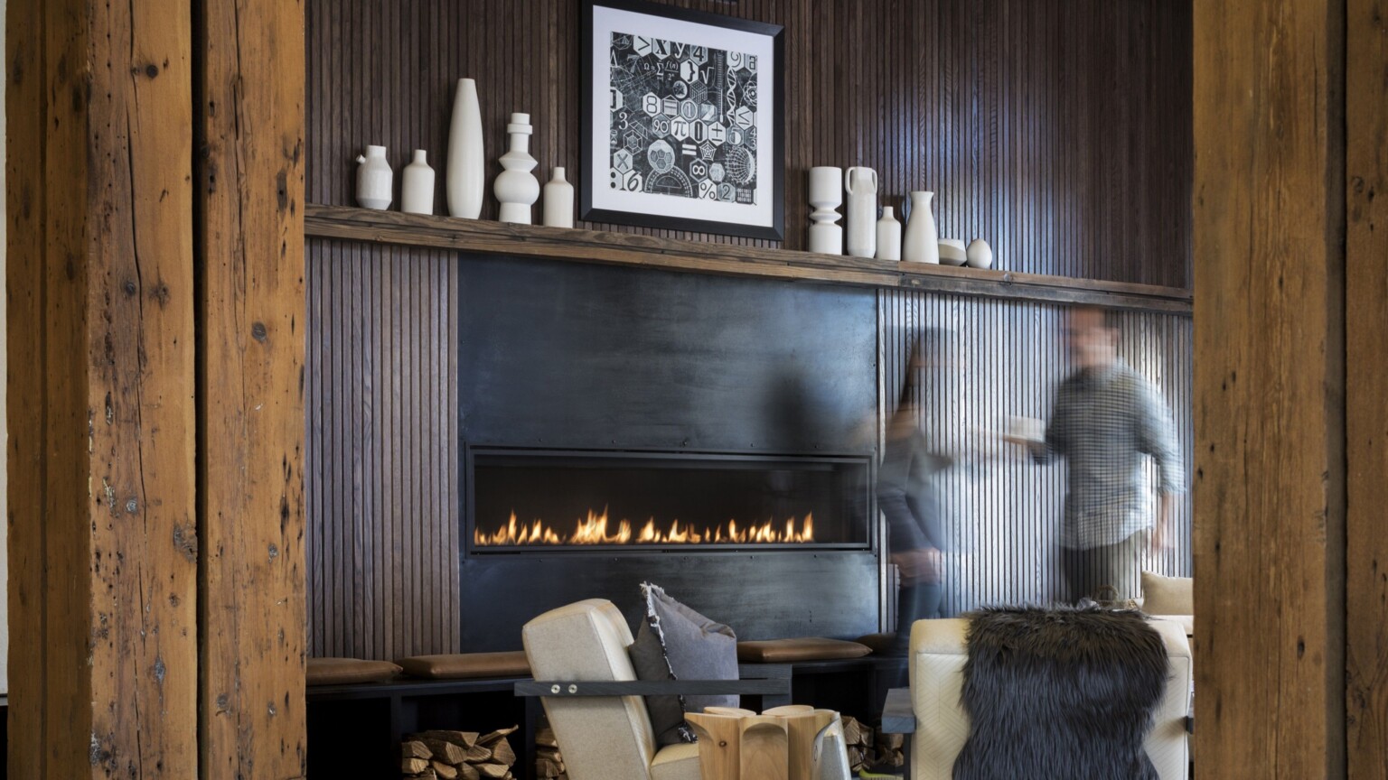 Fireplace elevated on black bench with under seat storage between wood pillars in bar with floor to ceiling windows; white ceramic vases line the fireplace mantle