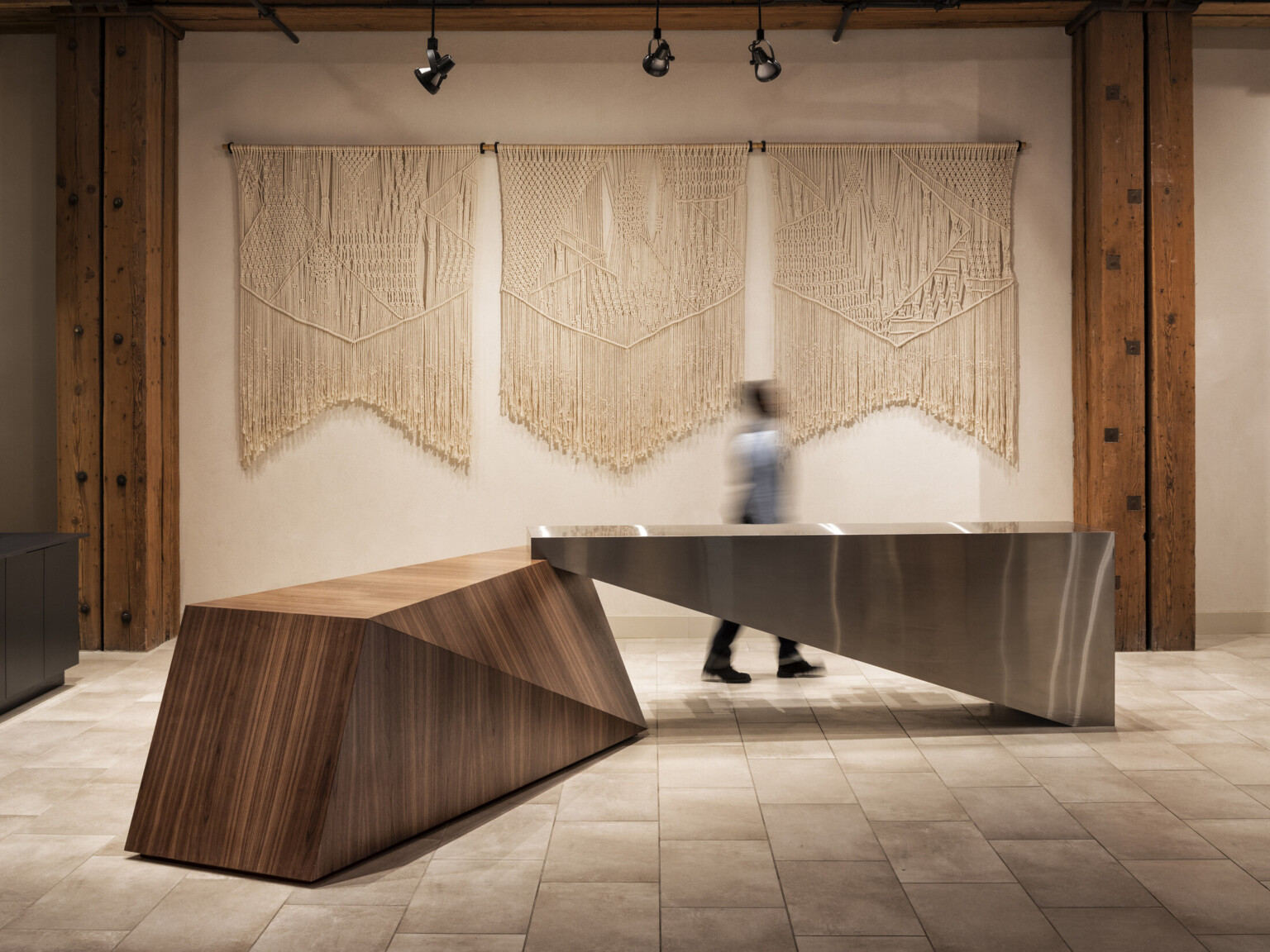 White wall with macrame wall hanging between wood pillars on tile floor with geometric wood and metal table at center