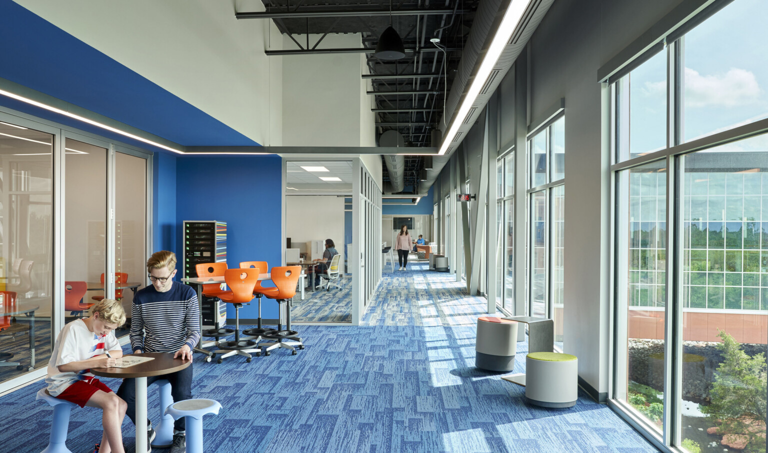 Blue carpeted hall with large windows on right wall. Left, recessed blue wall accent along glass wall looking into classes