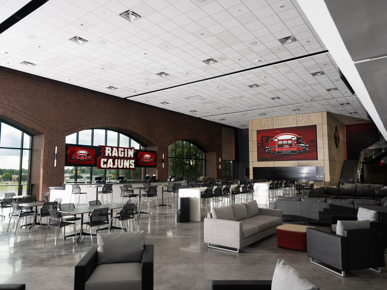 University of Louisiana Our Lady of Lourdes Stadium west bowl transformation; athletic lounging space with varied seating and bar space; red white and gray color palette with "Ragin Cajuns" sign on top of the bar