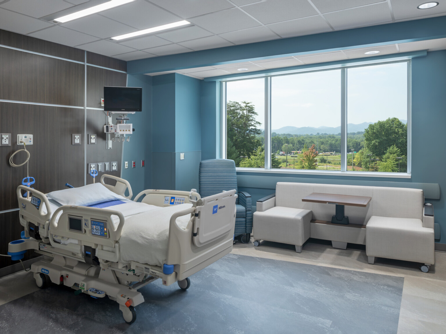 Modern hospital room with a bed, medical equipment, and a seating area by a large window