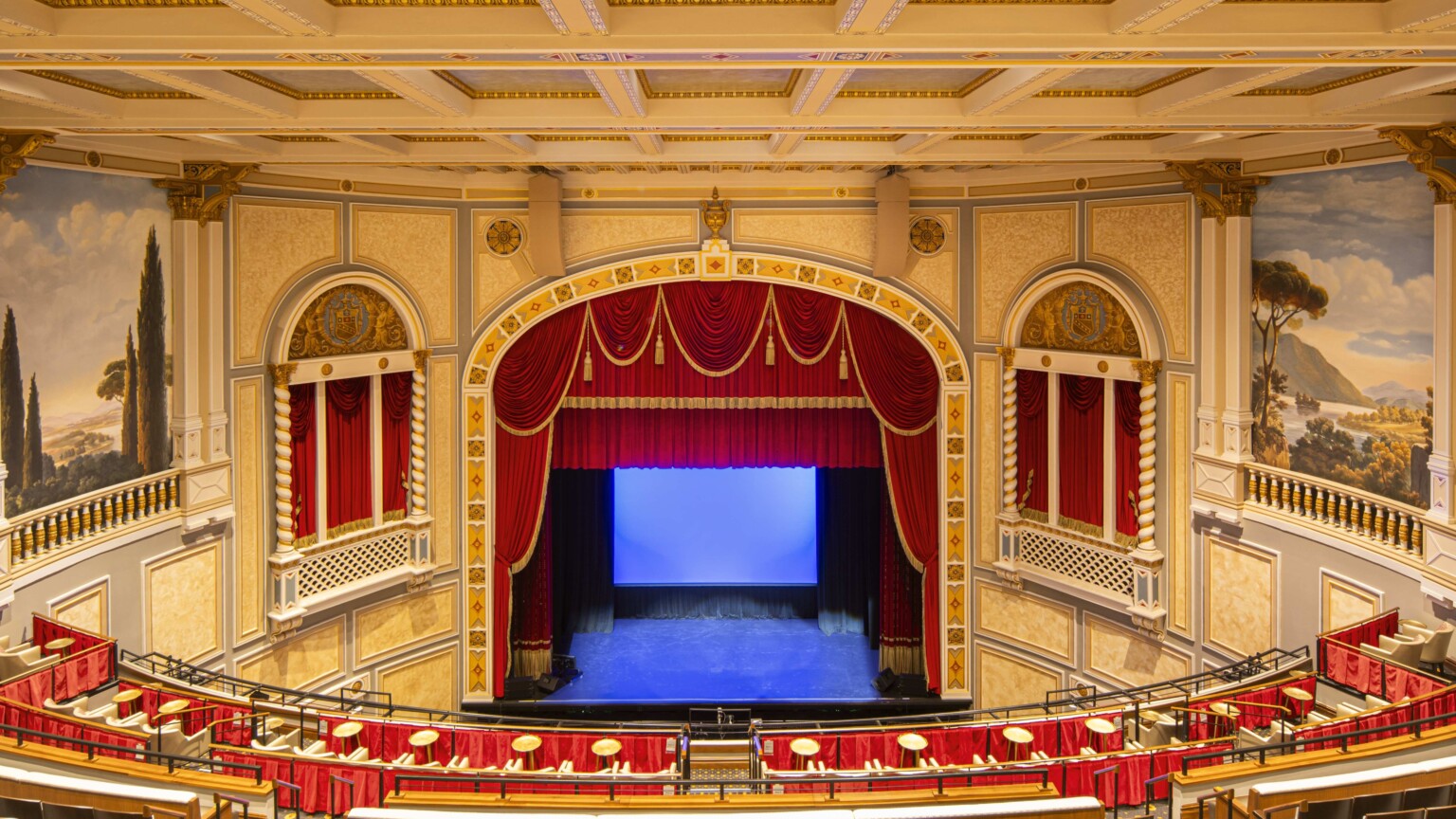 Interior of ornate theater with red velvet curtains, classical murals, and balcony seating; a private boxes with curtains