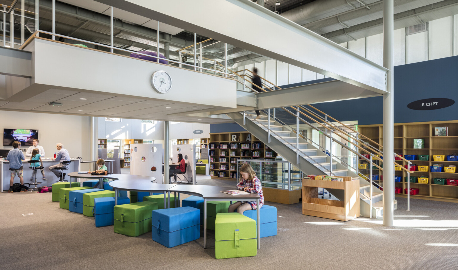 painted steel mezzanine with staircase over curved table with colorful soft seating next to bookshelf and clerestory windows