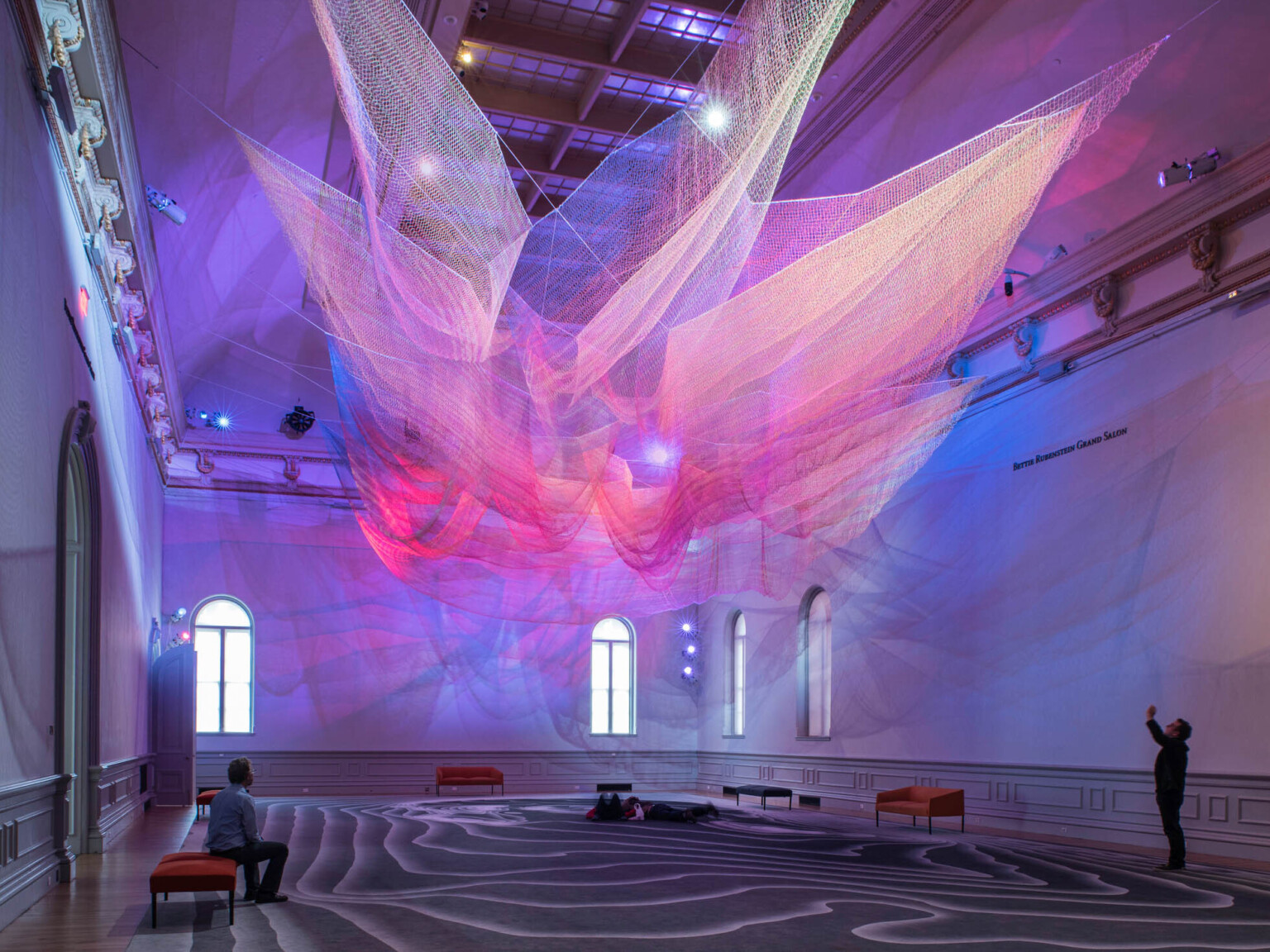 Colorful abstract art installation with pink and purple light on layers of sheer netting with shadows and light on white wall
