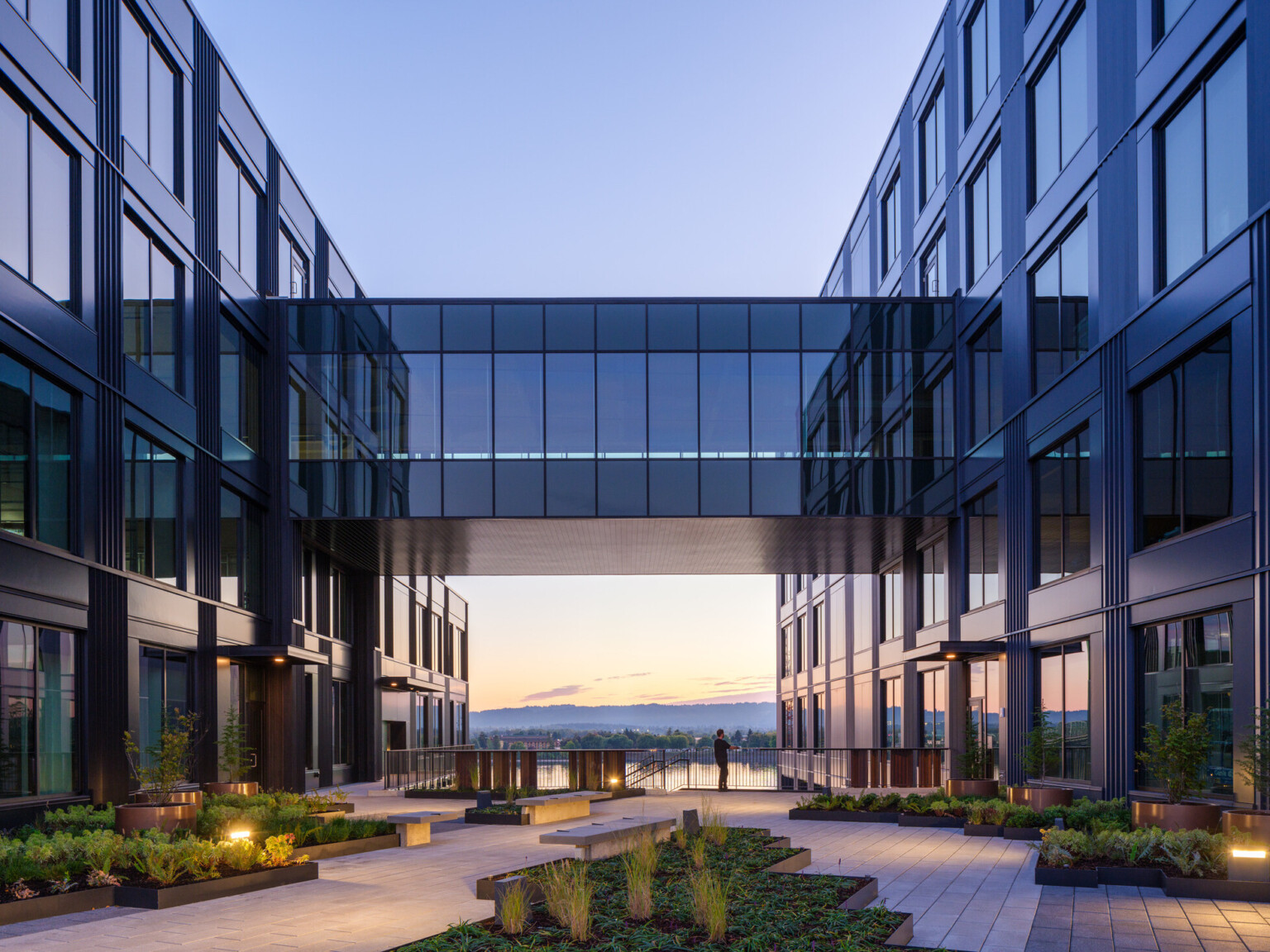night view of development on the water in vancouver, wa; mixed-use development with dark facade, elevated walkway, skyway connecting two buildings, courtyard for wellness