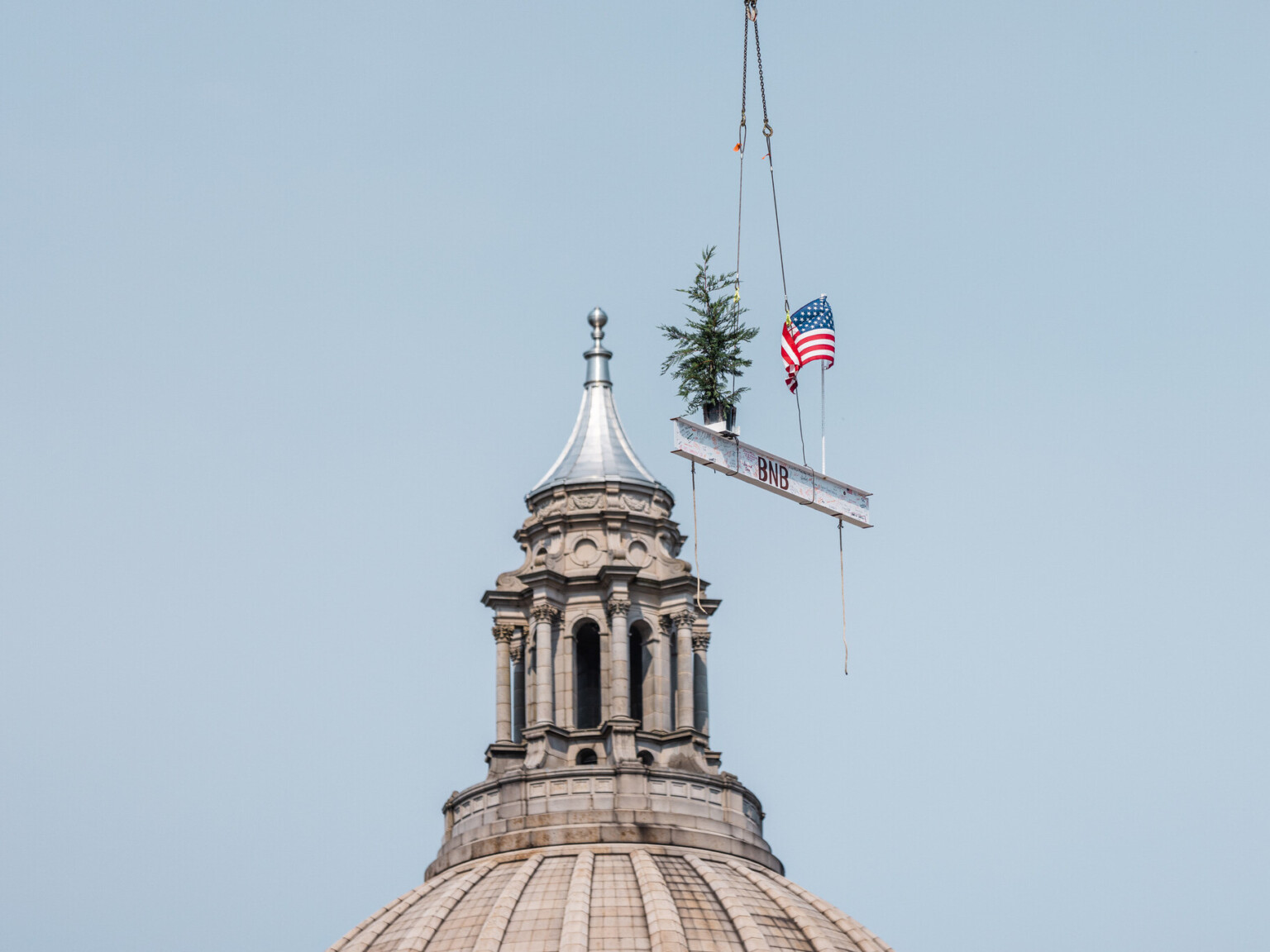 Helicopter flies steel beam bearing a potted cedar tree and the US flag past the Washington State Capitol dome on its way to the Pritchard Building. Photo Courtesy of Taras Yaremchuk/BNBuilders