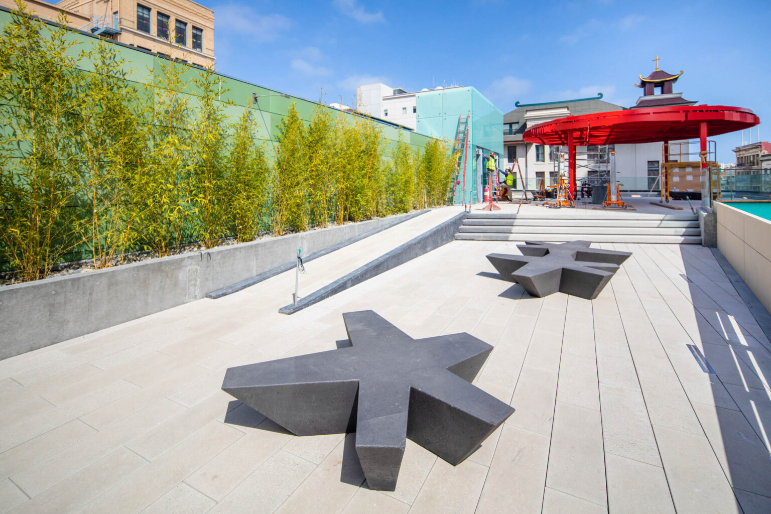 Organic shaped benches in walkway with ramp and steps to rooftop plaza with red canopy. Trees grow along green tile left wall
