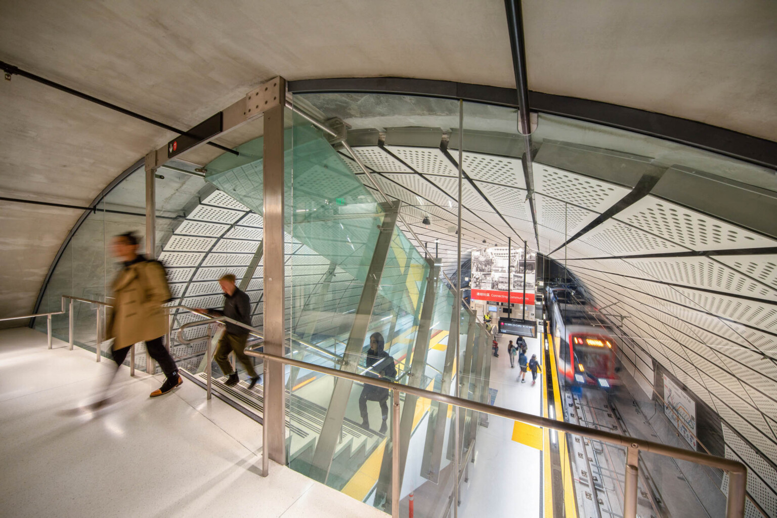 Rounded room with escalator to station below between 2 train tracks. White panel ceiling. Glass wall dividing tunnel
