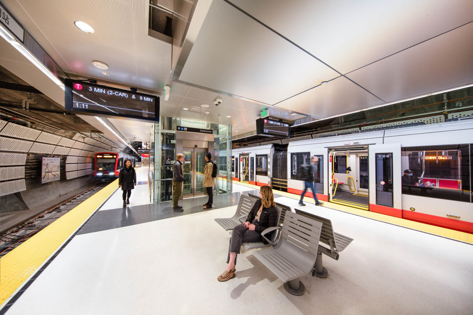 Rounded side walls of tunnel covered in perforated white panels. Hanging light beams along tracks beside screens, elevator at platform center