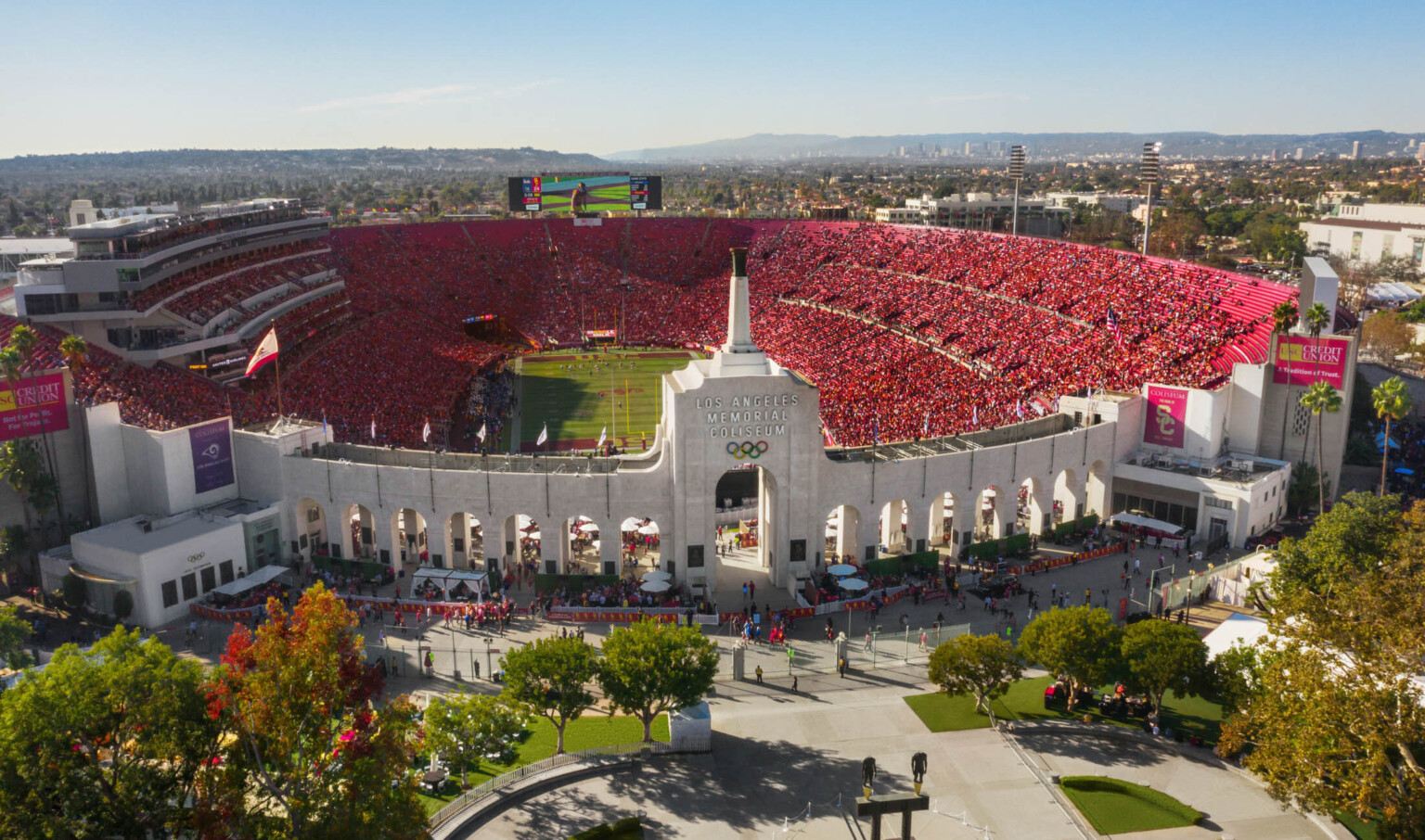 Front view of full stadium labeled Los Angeles Memorial Coliseum with Olympic Rings logo on central tower framed by archways