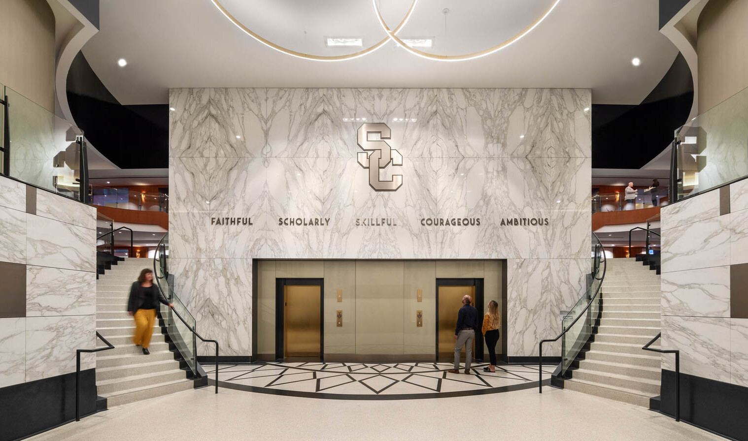 Marble side wall reading Founders Club frame curved double staircases and elevators in white room with round hanging lights