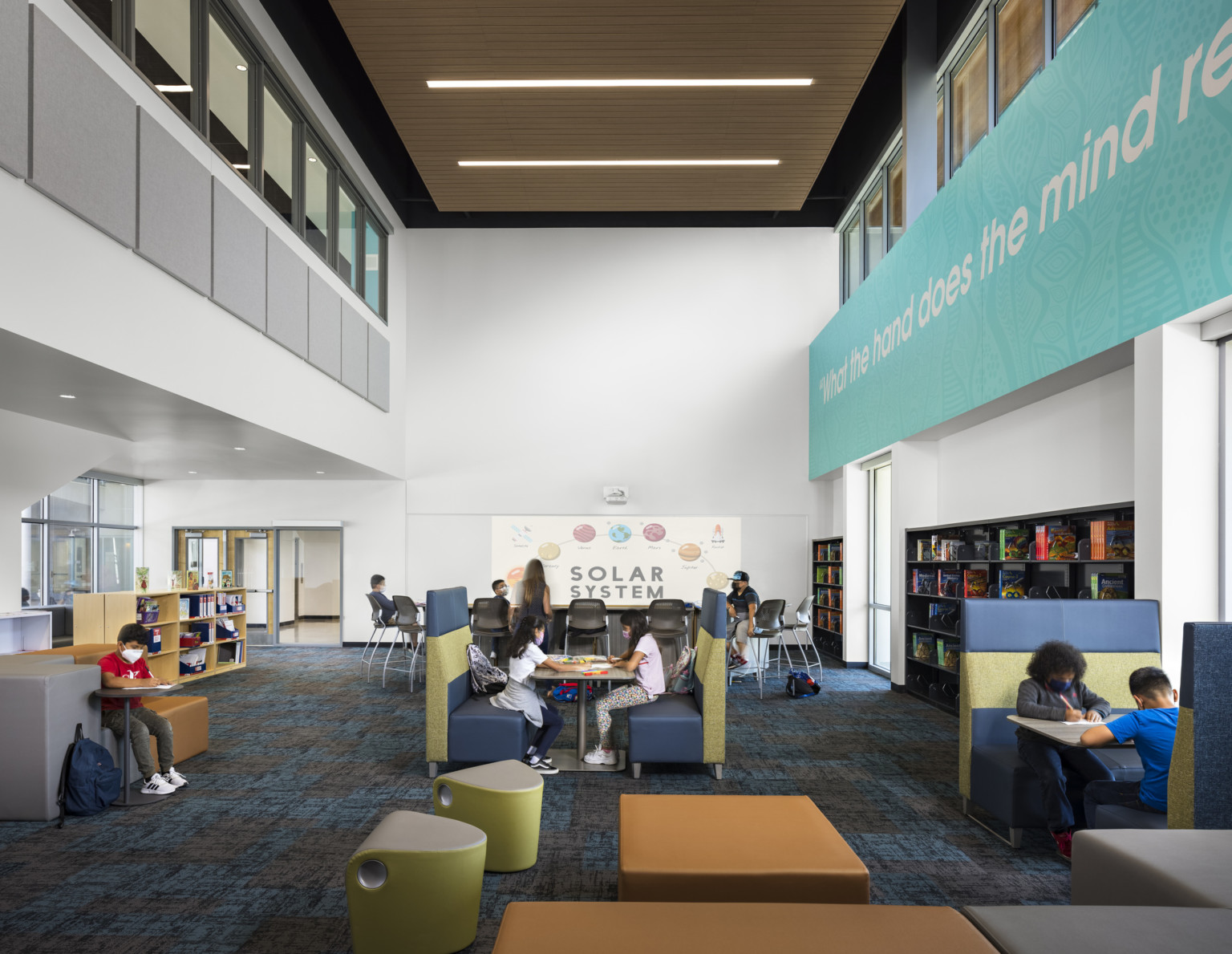 Double height seating area in Logan Memorial Educational Campus with shelves and large windows; school design with solar system hanging on the back wall; Interior windows above