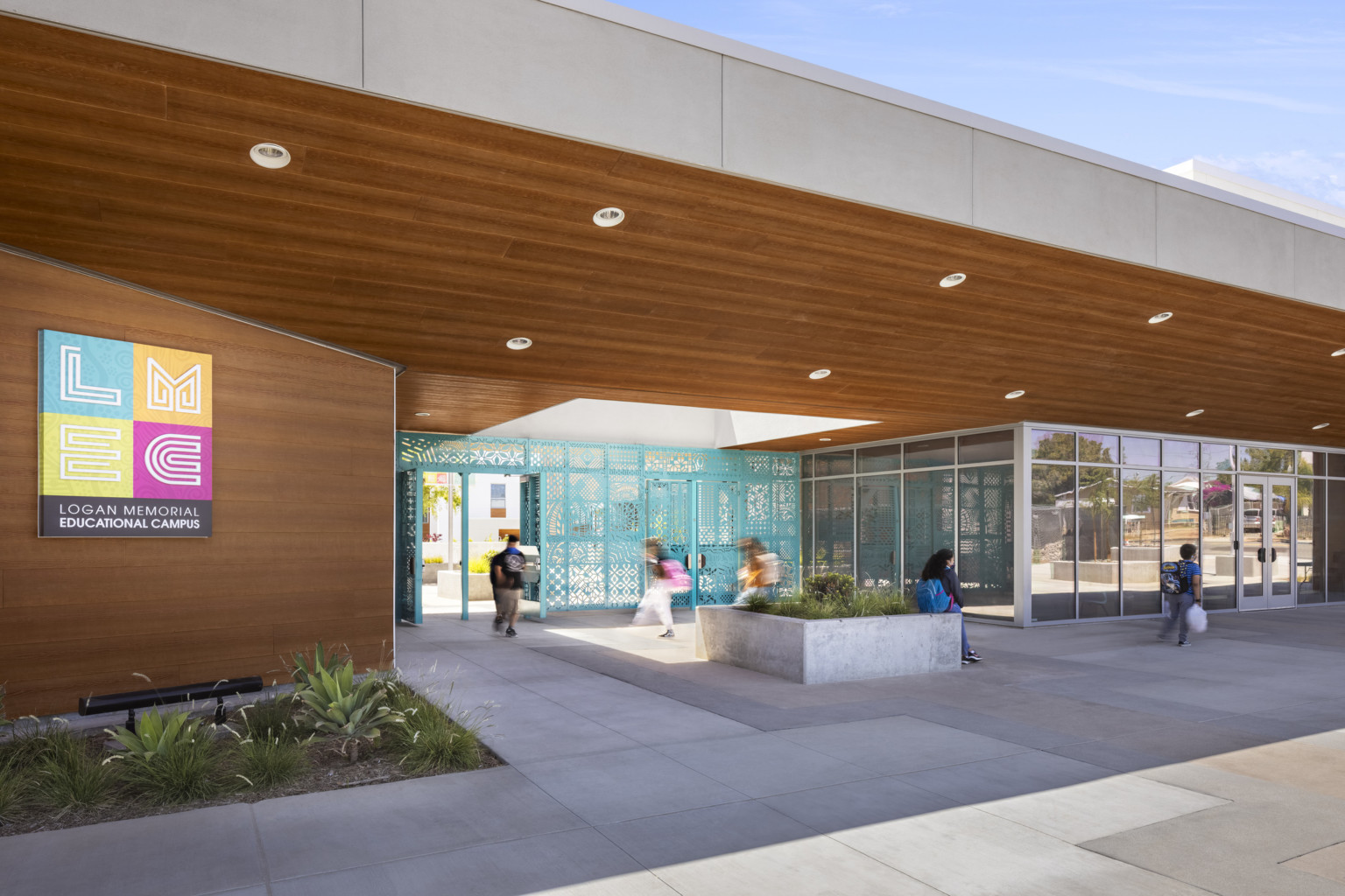 Entrance of Logan Memorial Educational Campus; school design elements with blue metal patterned gate recessed under a white overhang with wood panels underneath and a skylight at center