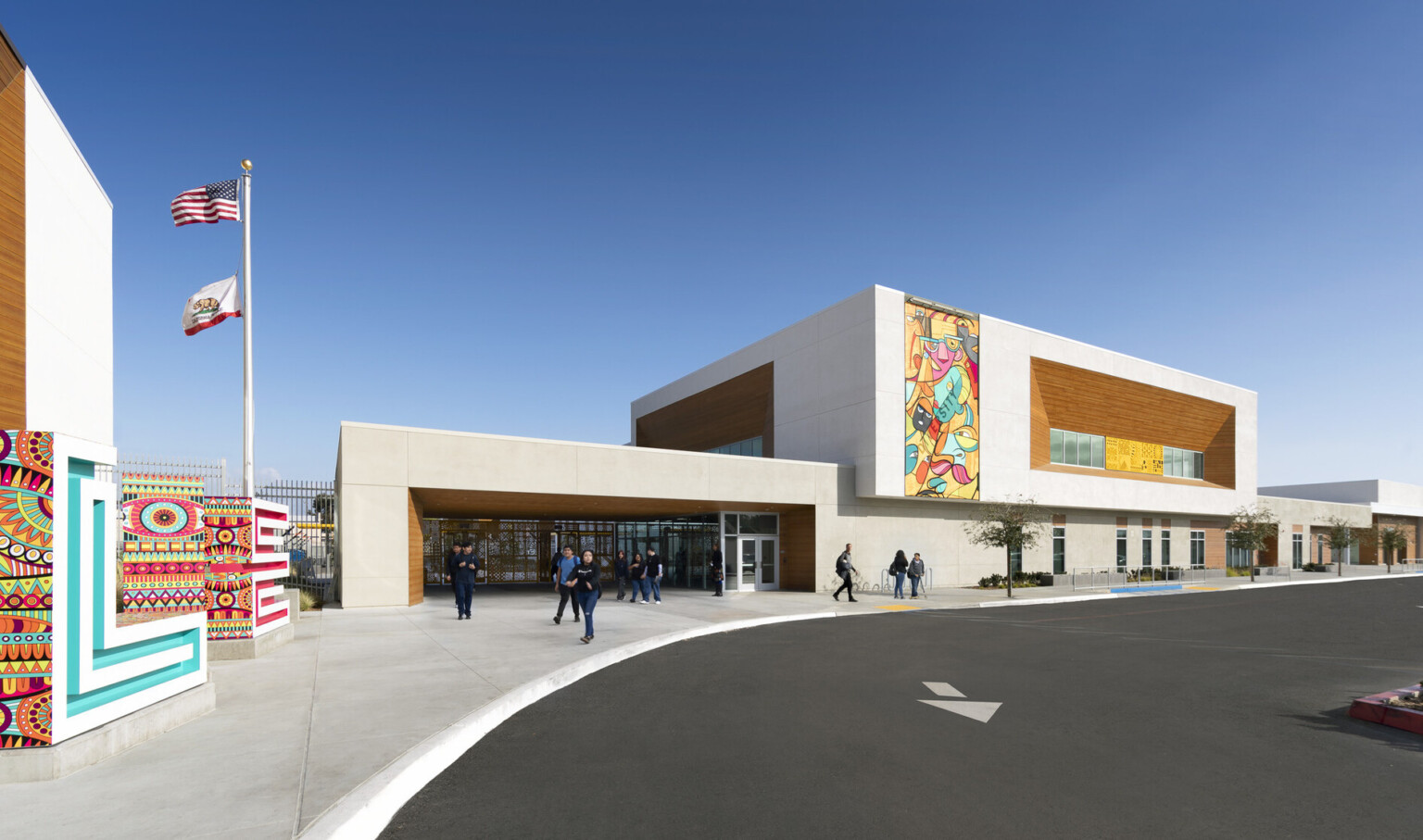 exterior view of Logan Memorial Education Campus entrance; parking lot; school design with brightly colored murals painted on two beige concrete and mass timber paneled façades, flagpole flying two flags