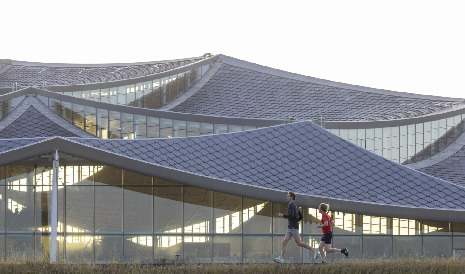Two people running in front of a building with an all glass facade and rectangular glazing units and a tiled sloping roof; google bay view