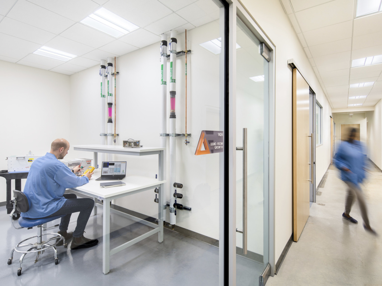 a glass room off a white hallway contains multilevel workspace and tubes connecting to wall. A man works with equipment