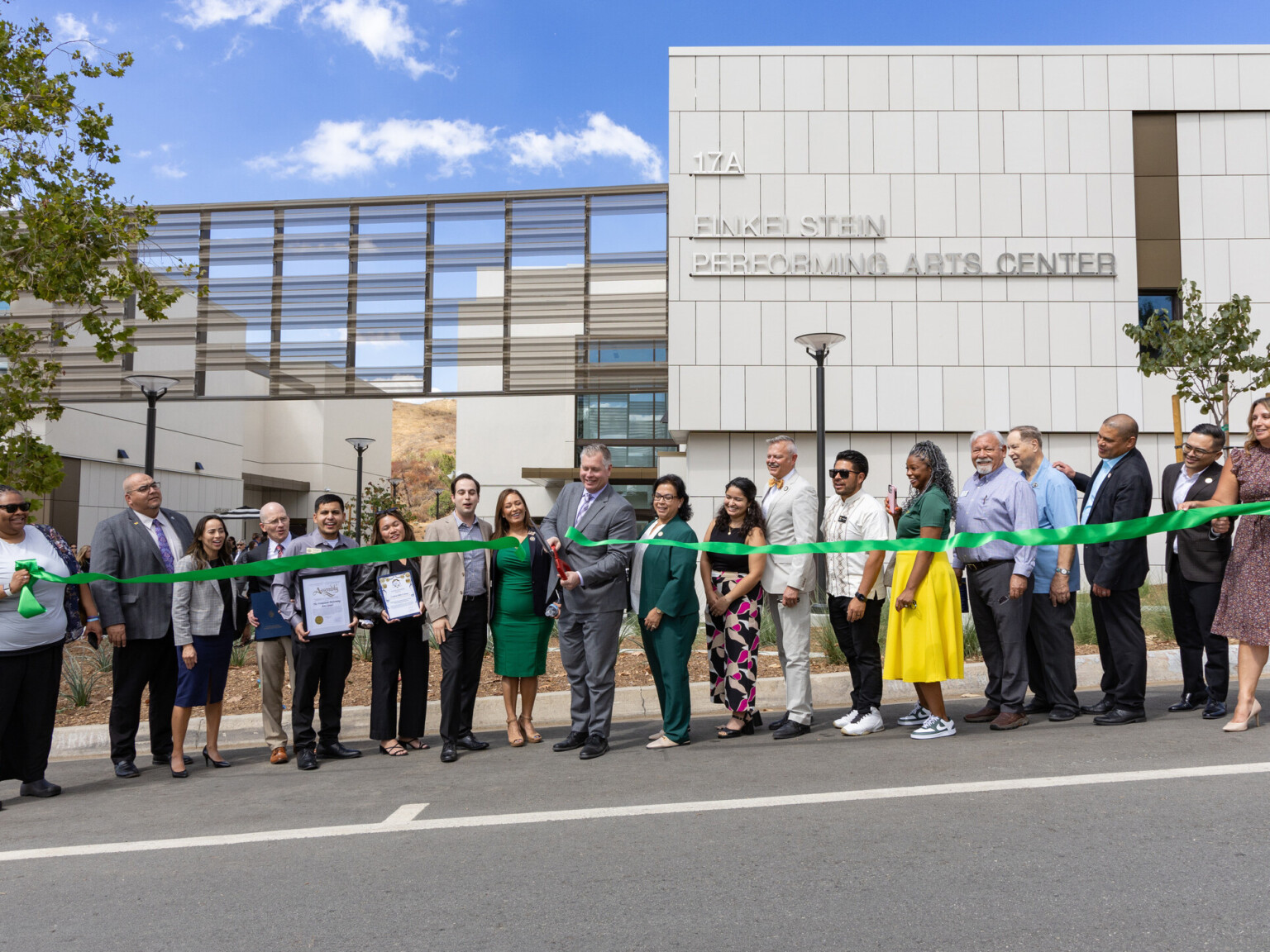 A group of people stands in front of a modern building. They are holding a long green ribbon, which is being cut by a person in the center using large scissors.