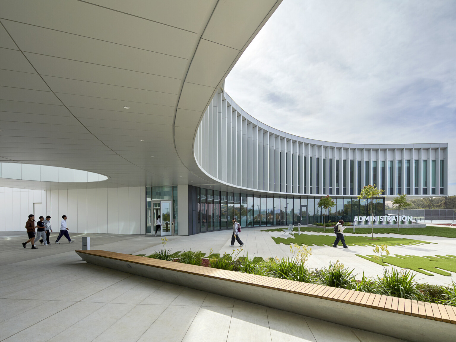 modern curved building with open green space and walking area beneath building; curved glass facade against a blue sky