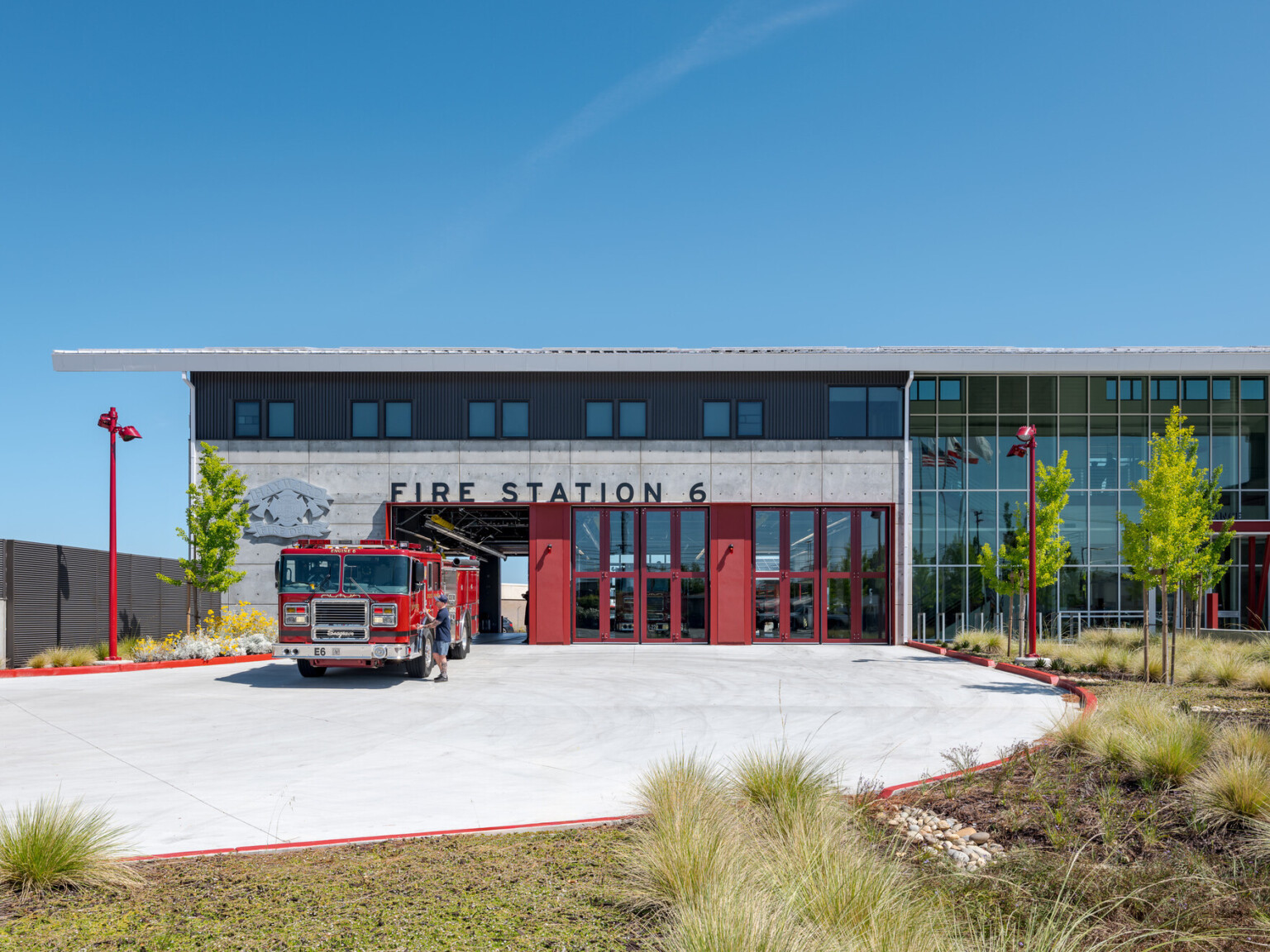 Exterior of white concrete a fire station with red accents. Words fire station 6 prominently displayed above the large glass doors with a fire truck parked at one of the bays.