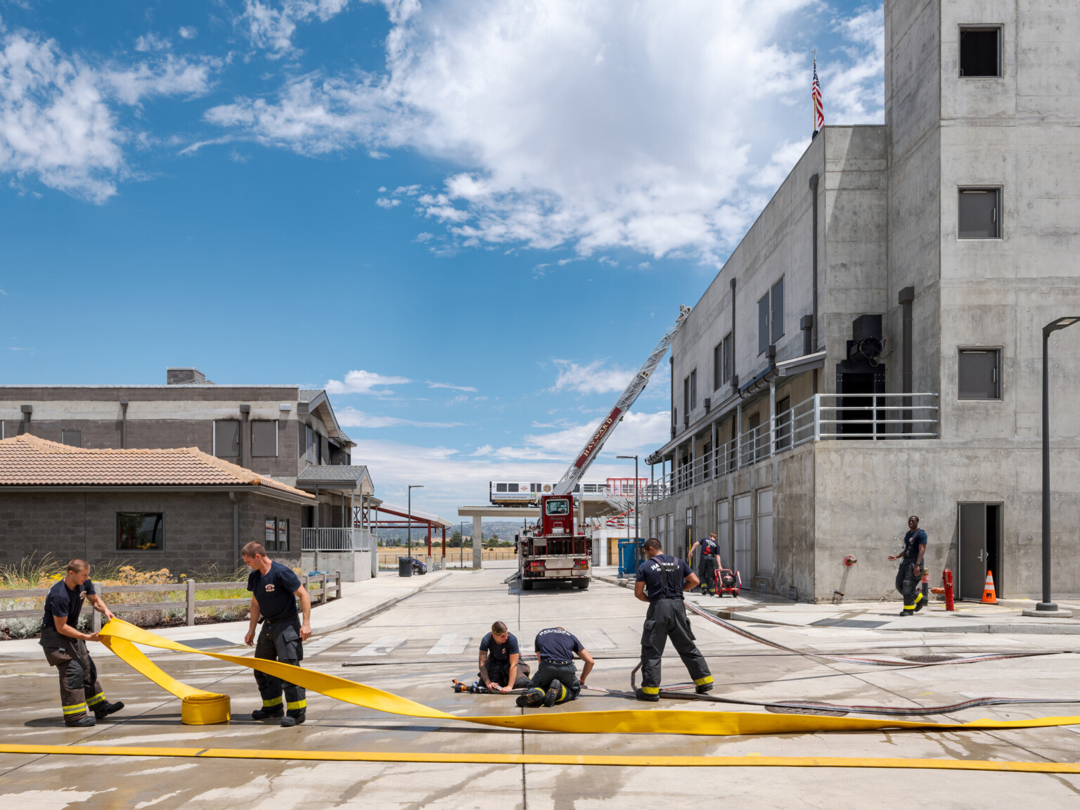 Fire fighters training outside a facility rolling up hoses with a fire truck with ladder extended in the background