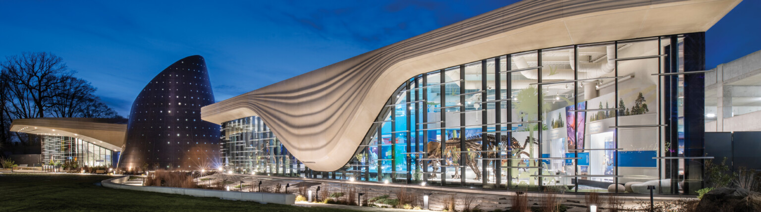 Nighttime image of a building with a white striped overhang that bows in center of right building section, an angled tube metal structure is at the building center
