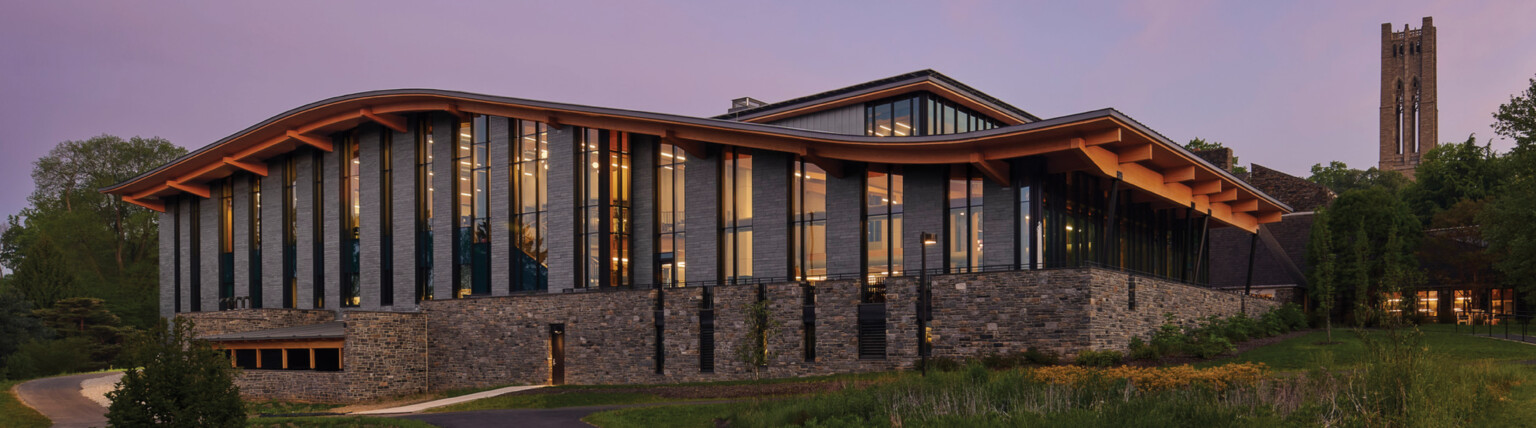 Stone and glass facade on university building with curving roof and overhang illuminated from within in the evening, mass timber architecture, what is mass timber construction, swarthmore