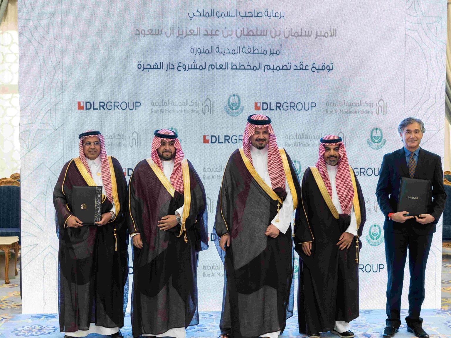 Five men standing in front of a white projection with DLR Group and Rua Al Madinah Holding across the background; two men on the ends holding portfolios with contracts