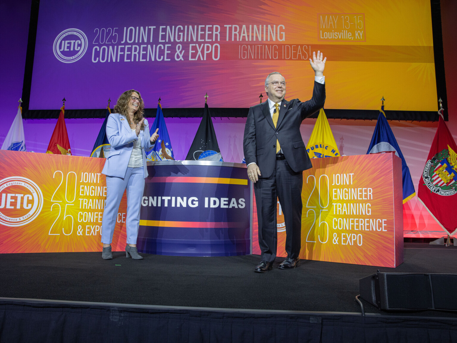 Michael Huffstetler elected to the Society of American Military Engineers; two people on stage celebrating the election with military flags and colorful banners behind them